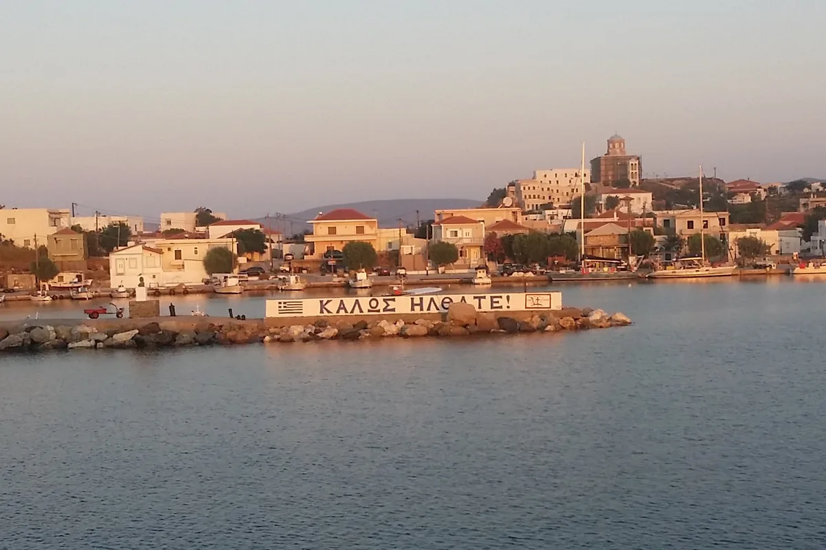 A Greek harbour village at golden hour — a painted sign on the breakwater reads ΚΑΛΩΣ ΗΛΘΑΤΕ, Welcome!